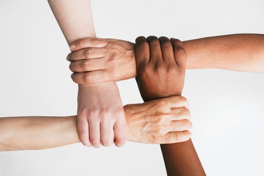 Four persons are using their right hands to make a square knot  by each hand holding the other persons wrist. The viewer sees the human knot from above. The ethnicity of the different hands implies persons of African, Asian, Hispanic, and Caucasian descent , 