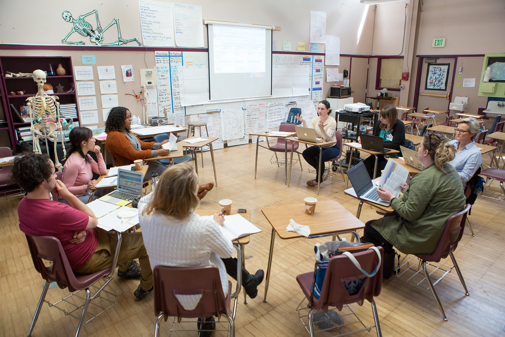 Eight teachers are sitting in school desks with cups of coffee and computers having a planning discussion.